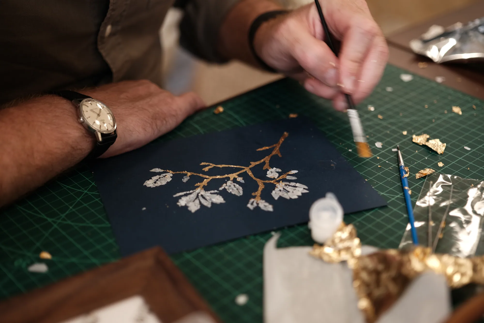 Hands applying gold leaf to a floral composition on a work table